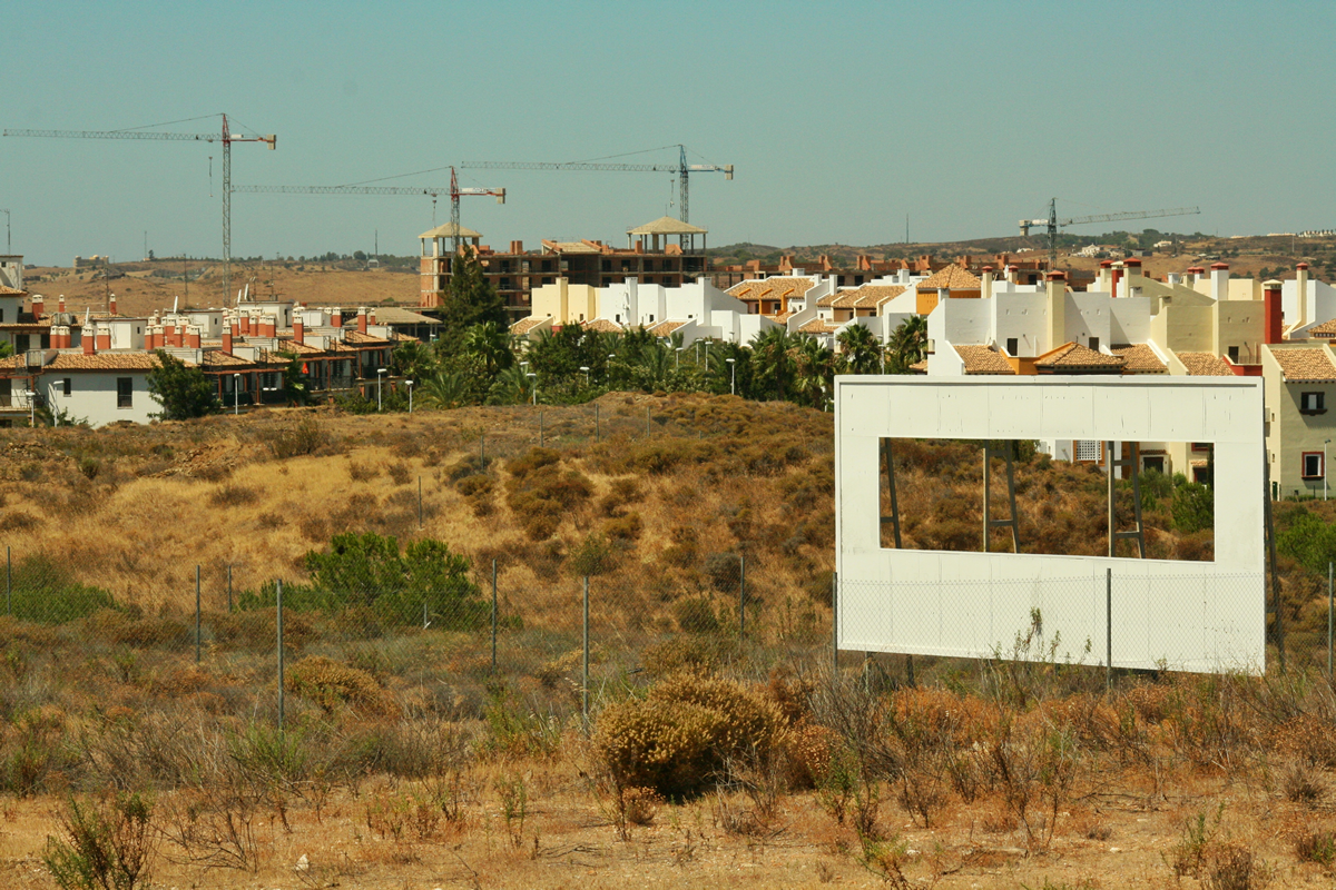 Transformaciones urbanas 2 (Costa Esuri &ndash; Ayamonte, Huelva)
&iquest;D&oacute;nde termina la ciudad y empieza el campo? La dificultad para encontrar una respuesta muestra que la distinci&oacute;n cl&aacute;sica entre lo urbano y lo rural ha perdido en muchas partes del mundo su significado. En los l&iacute;mites f&iacute;sicos (y conceptuales) de ambos dominios se localizan nuevos procesos de ocupaci&oacute;n espacial &mdash;denominados ciudad difusa, urbanizaci&oacute;n del campo, periurbanizaci&oacute;n...&mdash; surgidos al calor de un modelo econ&oacute;mico de desarrollo entregado al hormig&oacute;n y el ladrillo. Sin embargo, la resaca del boom inmobiliario nos ha dejado con los despojos de aquella borrachera: paisajes desordenados, fragmentados y carentes de identidad; enormes bolsas de suelo inacabado, residenciales medio abandonados, urbanizaciones "fantasma", etc. El ejemplo de la fotograf&iacute;a es paradigm&aacute;tico: se trata de Costa Esuri, un proyecto urban&iacute;stico y tur&iacute;stico junto a la desembocadura del r&iacute;o Guadiana de unas 400 ha (casi el doble de la superficie del propio n&uacute;cleo urbano de Ayamonte). Hoy en d&iacute;a este enorme complejo residencial, hotelero y de golf alterna algunas viviendas en uso con otras muchas en venta, a medio hacer o apenas proyectadas. Las ilusiones por convertir este rinc&oacute;n onubense en otro El Dorado se fueron desvaneciendo como la pintura de la valla publicitaria que lo anunciaba.